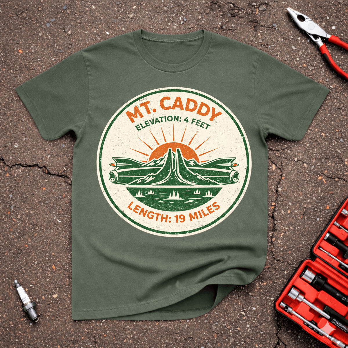 MT Caddy funny classic car T-shirt with retro mountain park badge, two long green classic cars, sunrise, and text reading Elevation 4 Feet Length 19 Miles on Military Green shirt.