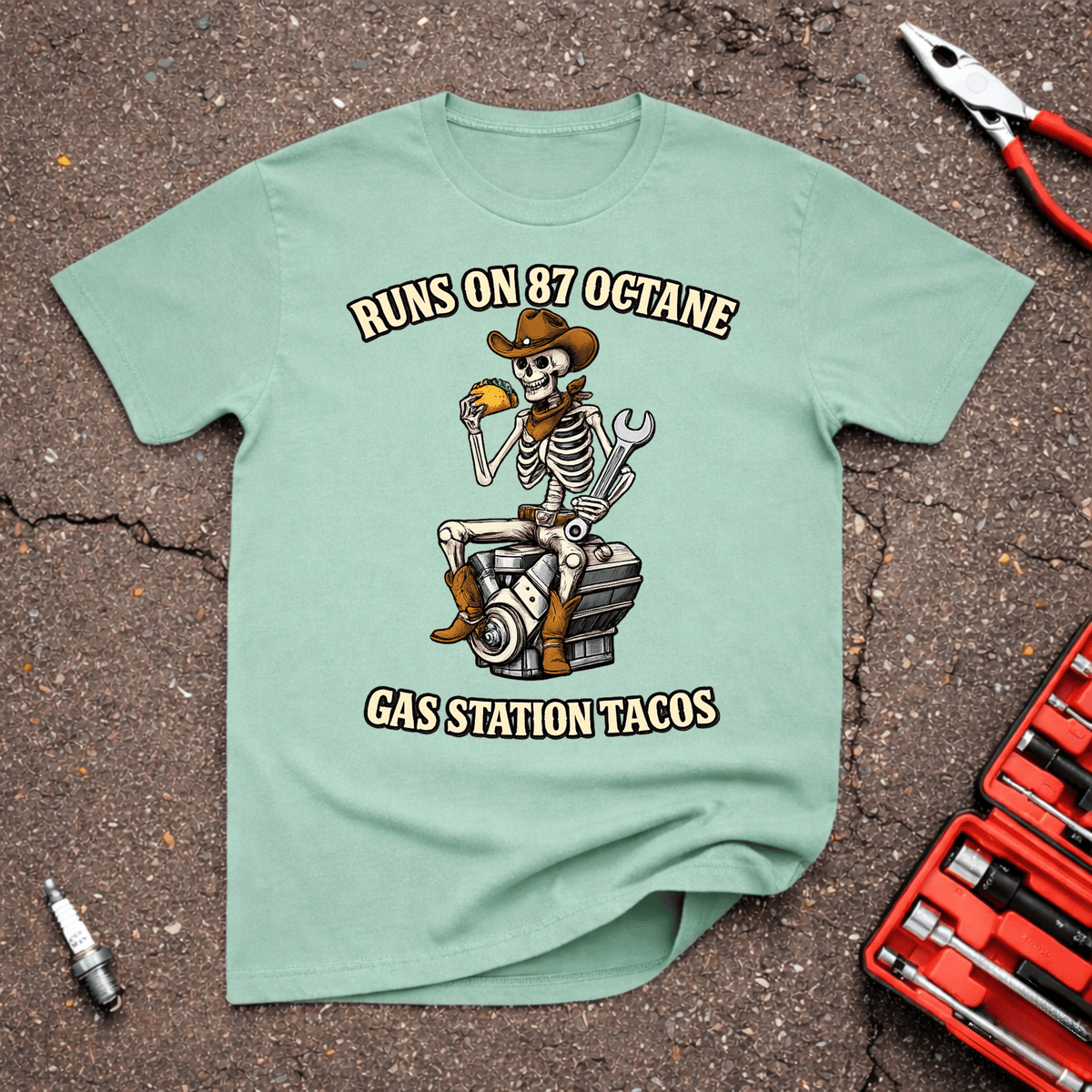 Tacos funny automotive T-shirt with cowboy skeleton eating a taco while sitting on an engine and Runs on 87 Octane Gas Station Tacos text on mint green.