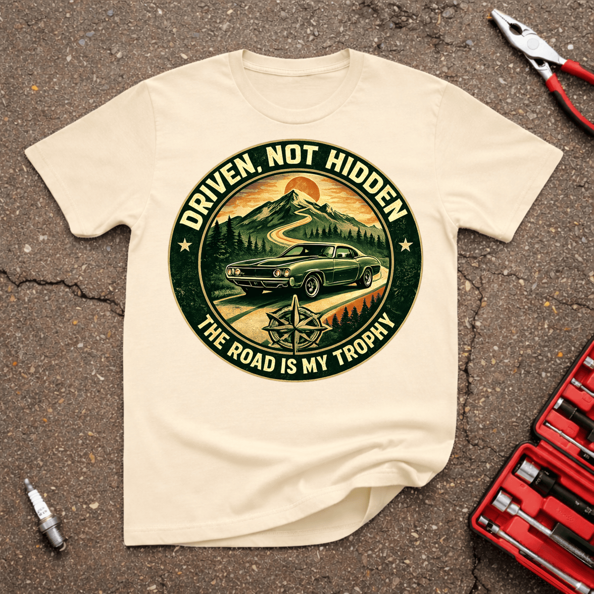 Road Is My Trophy automotive T-shirt with green classic car on winding mountain road, compass graphic, and Driven Not Hidden The Road Is My Trophy text on natural shirt.