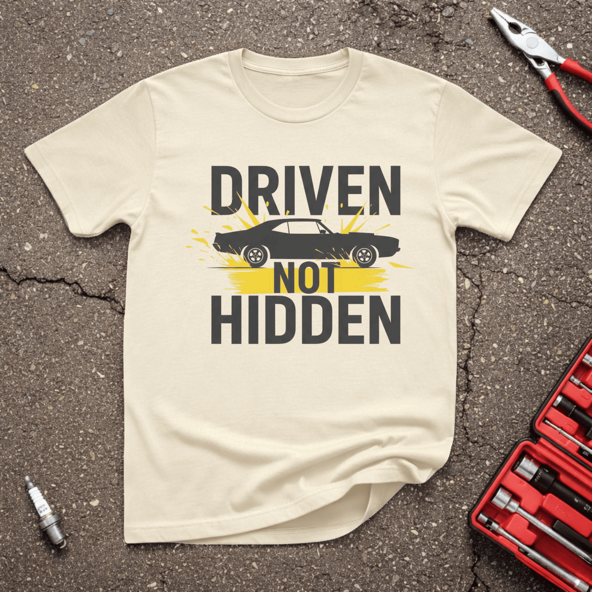 Driven Not Hidden sand automotive humor T-shirt with black classic muscle car silhouette, bold charcoal lettering, and yellow tire-smoke accents — GarageGiggles.com apparel.