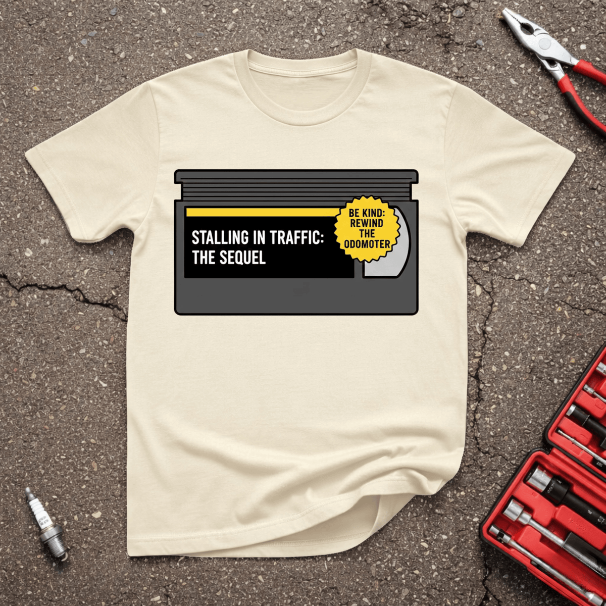 Be Kind Rewind funny automotive T-shirt with retro black video cassette reading Stalling In Traffic The Sequel and yellow sticker reading Be Kind Rewind The Odometer on Sand shirt.