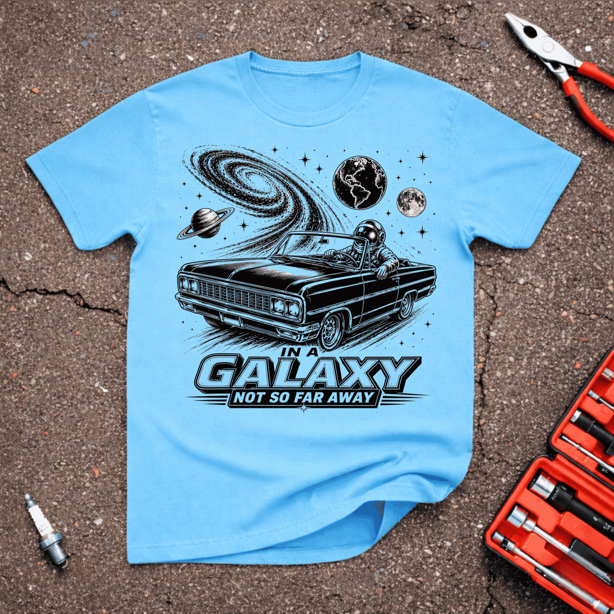 In A Galaxie funny automotive T-shirt with astronaut driving a black classic convertible beneath planets, moon, stars, and swirling galaxy on Sky Blue shirt.